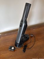 Small handheld Shark vacuum placed upright on charging base with power adapter cord visible, front view showing power button.