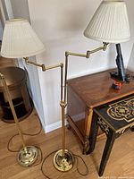 View of two gold-colored floor lamps with adjustable double arms and white pleated lamp shades, showing the full height and base of lamps.