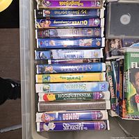 Photo showing a side view of VHS tapes stacked in a crate, titles visible include Disney classics like The Jungle Book, Oliver & Company, and others.