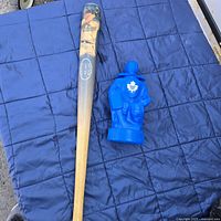Two items laid on a blue quilted surface: a wooden baseball bat with player image and a blue plastic bottle shaped like a hockey player.