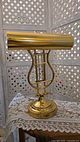 Photo showing the brass harp style banker’s lamp with a shiny brass cylindrical shade in a lowered position on a white lace cloth.