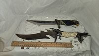 Full view of two decorative fantasy-style knives placed on white lace fabric with wooden ruler for size reference.