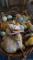 Basket filled with straw, pastel speckled eggs, and a decorative rabbit figurine with a floral hat.