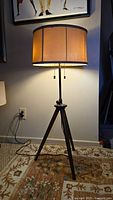 Front view of lamp lit, showing beige fabric drum shade with dark vertical accents, tripod wood-look metal legs, and pull chain switches.