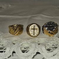 Photo of three men's costume rings arranged in a row on a white lace cloth background. The rings are gold tone with steel base and detailed designs including cross emblem and Harley-Davidson logo.
