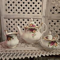 Royal Albert Country Roses teapot, sugar bowl, and creamer displayed together on lace tablecloth.