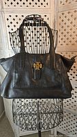 Front view of a black leather tote bag with shoulder straps showcasing a gold-tone Tory Burch logo on the front strap, displayed on a wire mannequin.