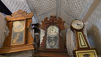 Three vintage clocks sitting on the floor against a lattice wall, showing their wooden carved frames and glass fronts with clock faces visible. The clock on the right has a long neck and decorated base.