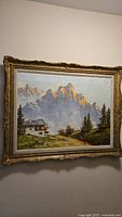Full view of vintage oil painting showing mountain peaks, cottage, and pine trees in ornate gold frame.