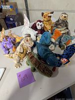 Photo showing 12 TY Beanie Babies plush toys on a table with visible TY tags, including various dinosaurs, dragons, and other creatures.