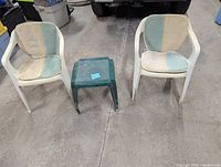 Front view of two white plastic chairs with blue and beige cushions and a small green plastic table between them on concrete floor.