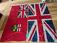 Photo showing two flags side by side on wooden floor: Ontario flag with shield and Union Jack flag