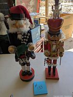 Two nutcrackers, Scottish-themed and toys/snowman-themed, standing on bases in well-lit area with boxes in background.