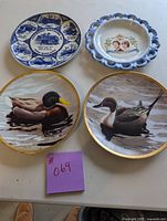Four collector plates on beige surface, showing all plates distinctly; includes blue and white covered bridge, Prince Charles and Lady Diana commemorative plate, and two duck plates.