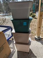Four empty plastic storage totes stacked outdoors in sunlight, with green Sterilite tote at the top and three white or gray totes below.