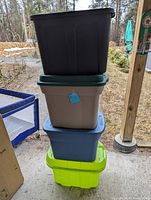 Stack of four plastic storage totes in green, blue, grey, and dark green colors, each with a lid, outdoors near a wooden post.