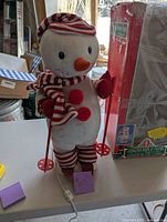 Full view of the snowman figure standing on skis with red and white striped winter accessories and ski poles on a table next to its box.