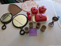 Photo showing the full assortment of kitchen items on table including two hand mirrors in black frames, wooden egg holders, metal iron holder, pot holder, red ceramic butter dish, salt and pepper shakers, condiment holder, and metal cup.