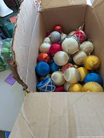 Open cardboard box filled with assorted old Christmas ball ornaments in colors red, white, blue, yellow, silver with various designs.