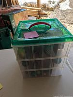 Green plastic 3-tier container with handle containing Christmas balls visible through transparent sides under green lid