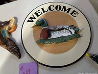 Close-up of the round Welcome sign featuring a mallard duck in the center, mounted on a white circular plaque with the word 'WELCOME'