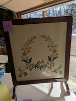 Frontal image showing wood-framed glass top with embroidered floral design on surface, standing upright.