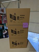 Three stacked cardboard boxes labeled 'Comfy Packages Hot Cups 16 oz 50 Sets' in a garage or storage area.