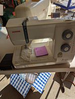 Front view of the Janome sewing machine with visible stitch selector dials and spool holder on top, placed on a table with blue checkered fabric beneath.