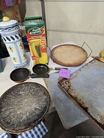 Various kitchen items shown on table including spaghetti holder with lemon graphics, Pasta Express box, two small fry pans, round pizza stone with stand, round mesh pizza screen, two rectangular cookie trays