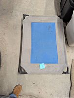 Top-down photo of pet cot showing blue cooling pad insert on elevated gray fabric frame on concrete floor.