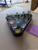 Nine pool ball themed shot glasses arranged in black triangular tray on outdoor surface, one missing from original set of ten.