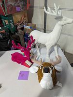 Three reindeer items on a white surface: white standing reindeer, pink plush reindeer, and brown plush reindeer head.