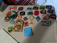 Top-down image showing 25 assorted embroidered scout patches, a green woven scout belt, and one round scout pin arranged on a table surface.