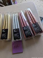 Five packs of tapered table candles laid out on a white table. Two packs are gold, two packs are red with glitter, and one pack is silver metallic colored. Each pack contains six candles, labeled Life at Home brand and measuring 10 inches tall.