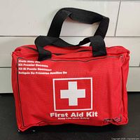 Closed red first aid kit bag with black handles and white cross symbol, showing front label in multiple languages.