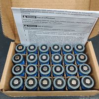 Open box showing 24 blue Amazon Basics CR123A lithium batteries arranged in rows with positive terminals facing up.