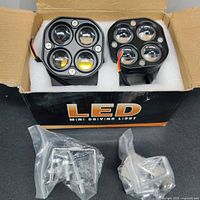 Two LED mini driving/work lights side by side in original box with mounting hardware in plastic bags in front.