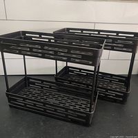 Two black, 2-tier pull-out spice racks made of plastic with metal supports and slider rails shown on a countertop.