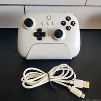 Front view of the white 8Bitdo Ultimate Bluetooth Controller seated in its charging dock with coiled USB cable in front.