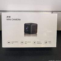 Front view of sealed white retail box with A18 Mini Camera label and image of small cube camera plus icons showing long battery life, WiFi phone app, motion detection, and night vision.