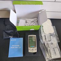 Open box showing all components including TENS unit, cables, electrode pads in packaging, use tips booklet, and carrying pouch.