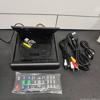 Top-loading DVD/CD player open showing disc tray with small USB stick inside, with remote control in plastic wrap, RCA cables, HDMI cable, and power cable laid out next to it.