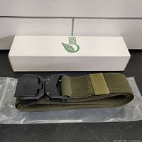 Full view of the rolled olive green tactical nylon belt with attached black powder coated quick release buckle, placed on a plastic packaging with a white branded box in the background.