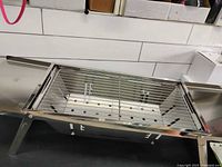 Stainless steel portable collapsible barbecue stove fully assembled on a black surface with four foldable legs extended, top view shows cooking grate and perforated base plate.
