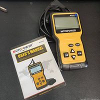 Yellow and black MOTOPOWER MP69033 OBD2 scanner next to user manual on black surface.
