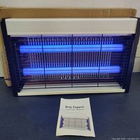 Bug zapper front view showing two glowing blue UV tubes inside a metal grid and the bug zapper instruction manual in front.