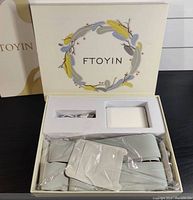 Open box showing the portable heating pad, attached belt straps, and accessories such as rechargeable battery pack and wiring, all neatly arranged in the original packaging.