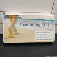 Box of grounding mat showing promotional text 'Reduces Inflammation, Reduction of Pain, Increased Energy, Stimulates Deeper Sleep' and image of walking barefoot on sand.