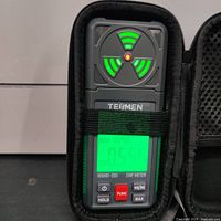 Front view of the TE8MEN Hound-200 EMF meter inside the black carrying case, showing digital display illuminated with a reading, buttons, and circular indicator with green lights.