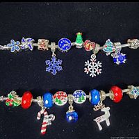Two silver-tone charm bracelets with assorted colorful winter and holiday-themed charms and beads displayed on black background.